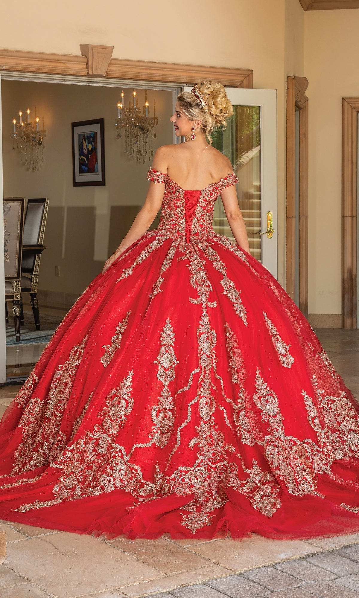 A woman models the Quinceanera Dress 1783 By Dancing Queen, featuring an off-the-shoulder design with gold lace embroidery, standing in a formal setting and showcasing its full, ornate skirt and train.